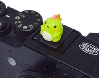 May include: A black digital camera with a green and orange dinosaur figurine on the hot shoe mount. The camera has a lens with a zoom ring and a black lens cap. The camera has the text "FUJIFILM X10" and "FUJINON LENS SYSTEM" printed on the top.