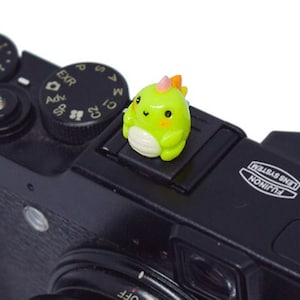 May include: A black digital camera with a green and orange dinosaur figurine on the hot shoe mount. The camera has a lens with a zoom ring and a black lens cap. The camera has the text "FUJIFILM X10" and "FUJINON LENS SYSTEM" printed on the top.