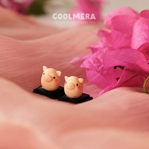 Può includere: Due piccole figurine di maialini rosa su basi nere. Le figurine sono sedute su una superficie di tessuto rosa. Lo sfondo è sfocato e presenta fiori rosa.