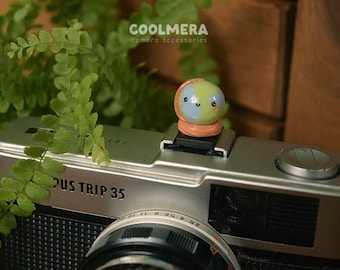 May include: A vintage camera with a light blue and green clay figurine of a planet with a smiling face on top of the lens. The camera is silver and black with the words "US TRIP 35" printed on the front.