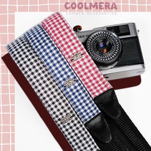 May include: Three camera straps with a black and white gingham pattern, a red and white gingham pattern, and a blue and white gingham pattern. The straps are attached to a black camera strap with silver buckles. The straps are labeled "COOLMERA camera accessories" and "STRIPE CAMERA STRAP".