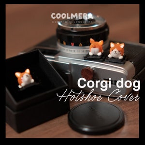 May include: A silver camera with a black lens and a brown wooden knob. Two orange and white corgi dog hot shoe covers are attached to the camera. The text "Corgi dog Hot shoe Cover" is written in black cursive on a wooden surface.
