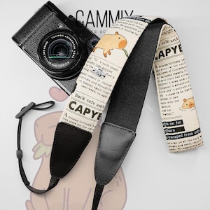 May include: A black camera with a lens is paired with a camera strap. The strap features a beige newspaper print with a capybara illustration and the text "CAPYE". The strap has black leather ends and a black adjustable cord.