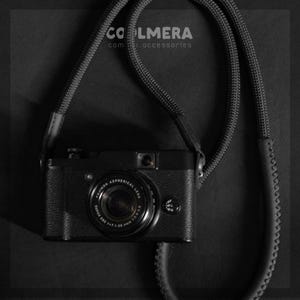 Puede incluir: Una cámara negra con una correa de cámara negra. La cámara tiene una lente negra con el texto "FUJINON ASPHERICAL LENS SUPER EBC 1:1.7-2.8 15-60mm" impreso en ella. La correa tiene el texto "COOLMERA com accessories" impreso en ella.