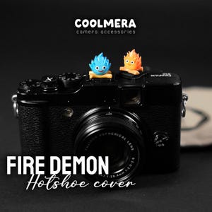 May include: A black camera with a blue and orange fire demon hot shoe cover. The camera has a lens with the text "FUJIFILM ASPHERICAL LENS 1:2.8 28.4mm 1:2.0-2.8" printed on it. The text "COOLMERA camera accessories" is printed above the camera.