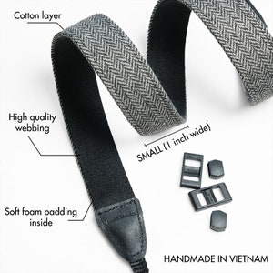 May include: A black and gray camera strap with a herringbone pattern. The strap is 1 inch wide and has soft foam padding on the inside. It includes two black plastic adjustable buckles and two black plastic strap connectors. The strap is handmade in Vietnam.
