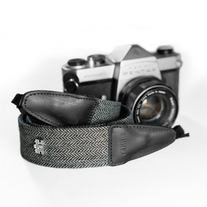 May include: A black and gray camera strap with a herringbone pattern and a black leather accent. The strap has the word "CAMIBOX" printed on it.