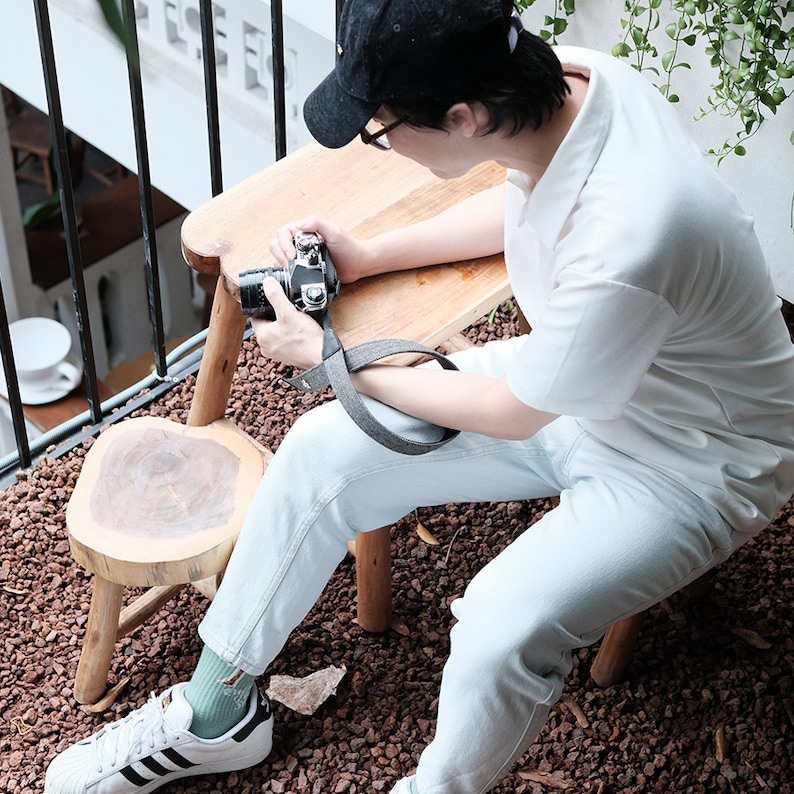May include: A person wearing a white polo shirt, blue jeans, and a black baseball cap sits on a wooden stool and holds a vintage camera. The person is sitting on a patio with a wooden table and a small stool.