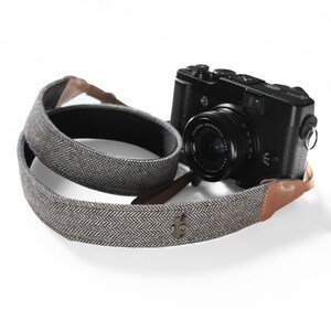 May include: A gray and white patterned camera strap with brown leather accents. The strap has a small metal logo with a design on it. A black camera with a lens is attached to the strap.