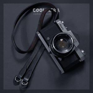 Puede incluir: Una cámara Kowa negra y plateada con una correa de cuero. La lente de la cámara es visible, junto con el texto "Kowa" y "1:1.8". La correa es negra con anillos de metal plateado. Las palabras "COOL" y "camera acces" son visibles.