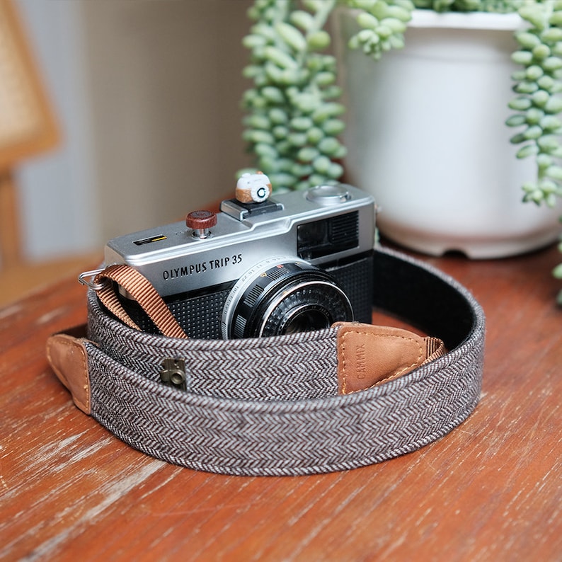 May include: A silver Olympus Trip 35 camera with a brown leather strap. The camera has a black lens and a brown leather strap with a herringbone pattern.