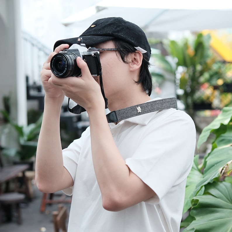 May include: A person wearing a black baseball cap and a white shirt is holding a black and silver camera with a black strap. The camera has a lens with a black cap.
