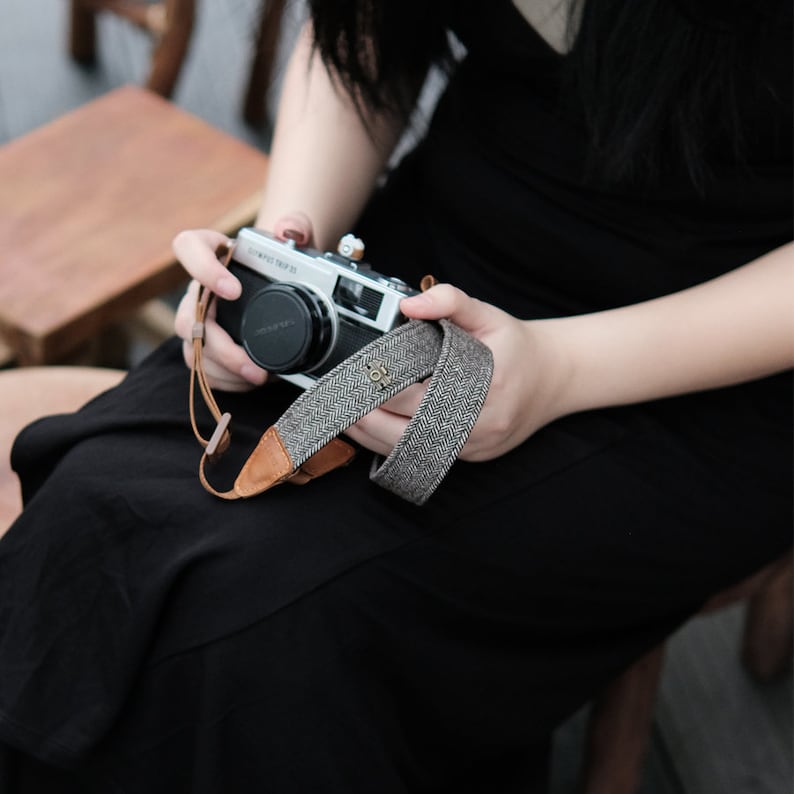 May include: A silver camera with a brown and gray camera strap. The strap has a herringbone pattern and a small metal charm.