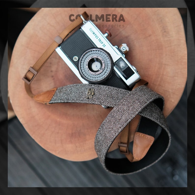 May include: A black and silver Olympus Trip 35 camera with a brown and gray tweed camera strap. The strap has a metal buckle with a design on it. The camera is sitting on a wooden surface.