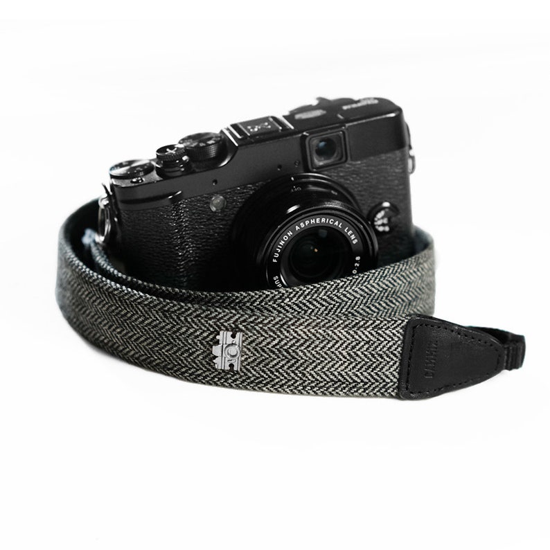 May include: A black camera with a black and white herringbone camera strap. The strap has a silver metal buckle with a logo.