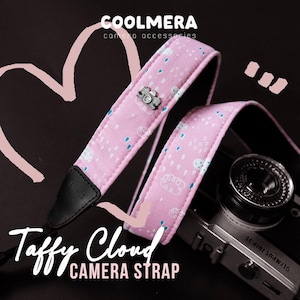 Puede incluir: Correa de cámara rosa con un patrón de nubes y gotas de lluvia, con un dije de cámara de metal. La correa está etiquetada como "Taffy Cloud CAMERA STRAP" y está diseñada para accesorios de cámara. La marca "COOLMERA camera accessories" también es visible.