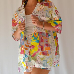 Puede incluir: Una camisa blanca de botones y pantalones cortos a juego con un estampado abstracto de colores. El estampado presenta formas geométricas, remolinos y puntos en tonos de rosa, amarillo, azul y verde.