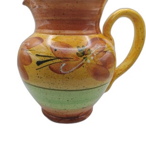 May include: A ceramic pitcher featuring a speckled finish with horizontal bands of brown, yellow, and green. The body is decorated with hand-painted orange floral motifs and black accents. It includes a curved handle and a wide pouring spout.