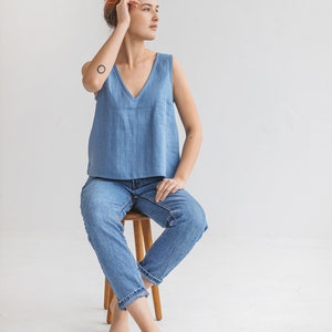 May include: A woman wearing a light blue sleeveless top and blue jeans sits on a wooden stool. She has red hair and is looking to the right.