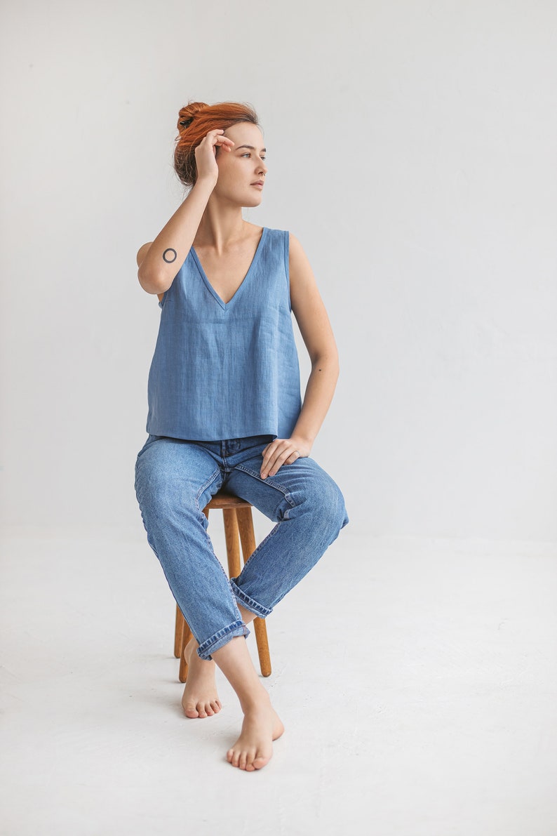 May include: A woman wearing a light blue sleeveless top and blue jeans sits on a wooden stool. She has red hair and is looking to the right.