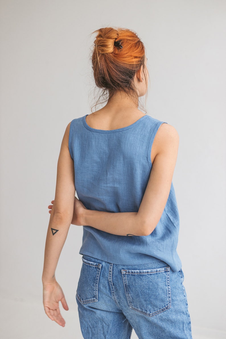 May include: A woman wearing a light blue sleeveless linen top and blue denim jeans. Her hair is pulled back in a bun and she has a small triangle tattoo on her arm.