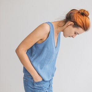 May include: A woman wearing a light blue sleeveless top and blue jeans. She is standing in front of a white wall.