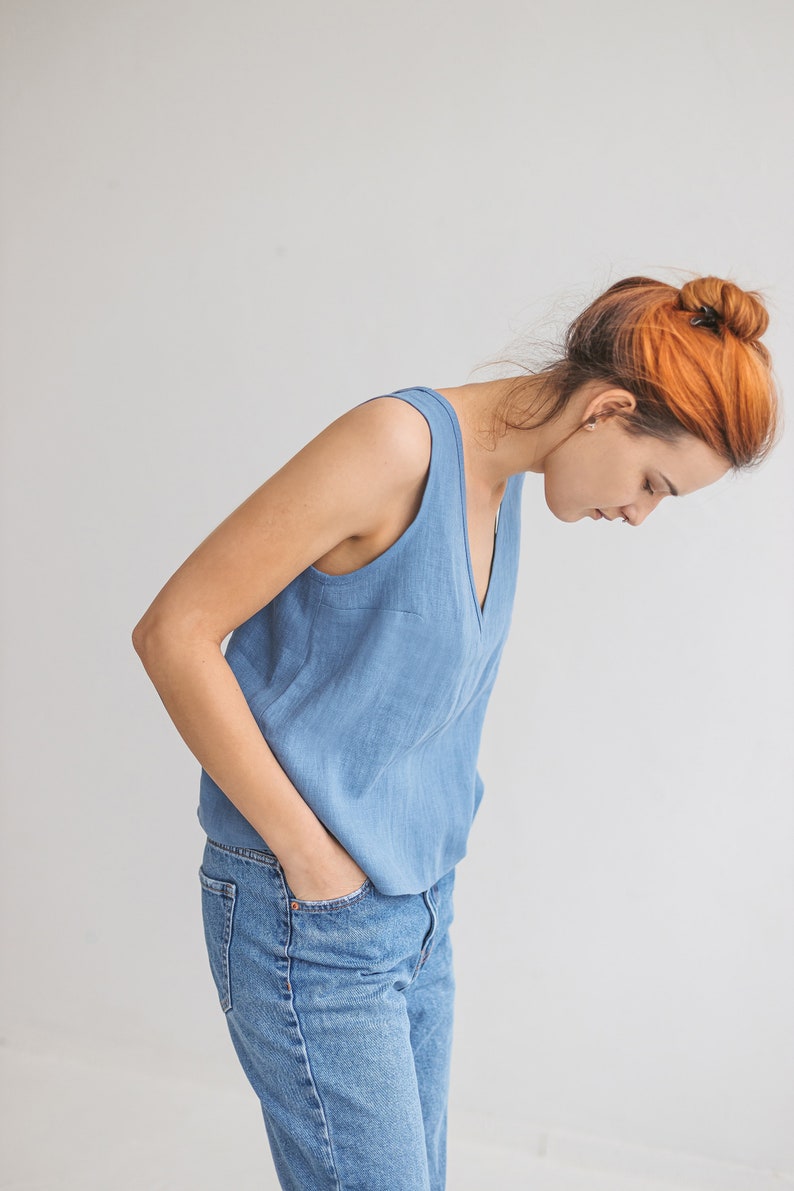 May include: A woman wearing a light blue sleeveless top and blue jeans. She is standing in front of a white wall.