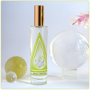 Può includere: Un flacone di vetro trasparente con un tappo dorato e un'etichetta verde e bianca che recita "ROSA CRISTICA". Il flacone è appoggiato su una superficie bianca con una sfera di vetro trasparente e un globo giallo.
