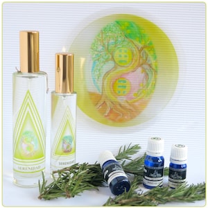 May include: Two glass spray bottles with gold caps and labels that say "SERENIDAD" in a yellow triangle. The bottles are on a white surface with green rosemary sprigs. Three small glass bottles with blue liquid and white caps are also on the surface. The labels on the small bottles say "LAVANDA", "PINO SILVESTRE", and "ANGELICA". A round, yellow, and pink plate with a painted tree design is in the background.