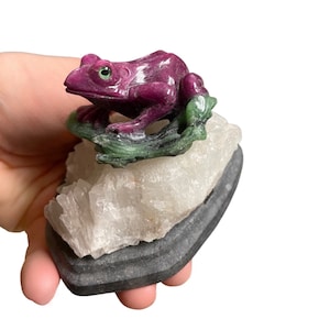 Puede incluir: Una figurita de rana de rubí tallada posada sobre una hoja de jade verde, sentada sobre un cúmulo de cristal de cuarzo blanco. El cúmulo de cristal está montado sobre una base negra.