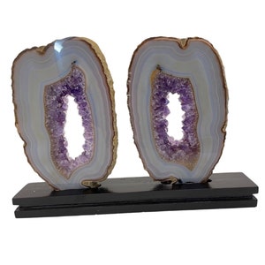 May include: Two agate slices with amethyst crystals in the center, mounted on a black wooden base. The agate is a light blue and white color with brown edges. The amethyst crystals are a deep purple color.