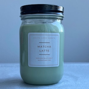 May include: A jar candle with a black lid and a label that reads "MATCHA LATTE." The candle wax is a light green color. The label also says "South Loop Scents" and describes the scent as earthy green tea with notes of vanilla and sweet cream.