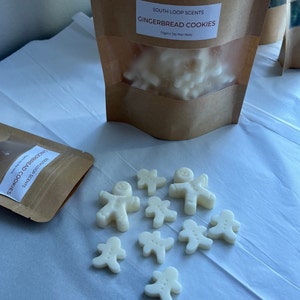 May include: A brown paper bag with a label that reads "South Loop Scents Gingerbread Cookies Organic Soy Wax Melts". The bag contains white wax melts shaped like gingerbread men. There are also several gingerbread man wax melts scattered on a white surface.