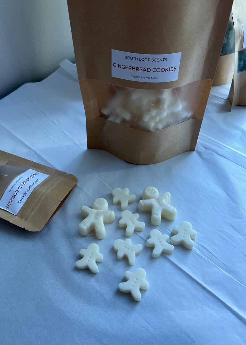May include: A brown paper bag with a label that reads "South Loop Scents Gingerbread Cookies Organic Soy Wax Melts". The bag contains white wax melts shaped like gingerbread men. There are also several gingerbread man wax melts scattered on a white surface.
