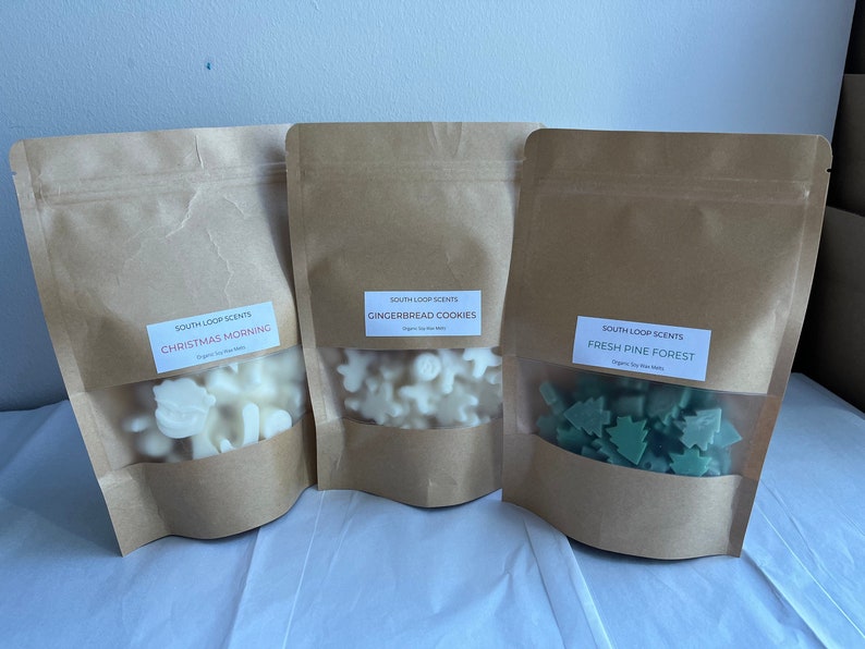 May include: Three brown paper bags with clear windows, each containing wax melts in different shapes and colours. The bags are labelled with the text "SOUTH LOOP SCENTS" and the scent names: "CHRISTMAS MORNING", "GINGERBREAD COOKIES", and "FRESH PINE FOREST".
