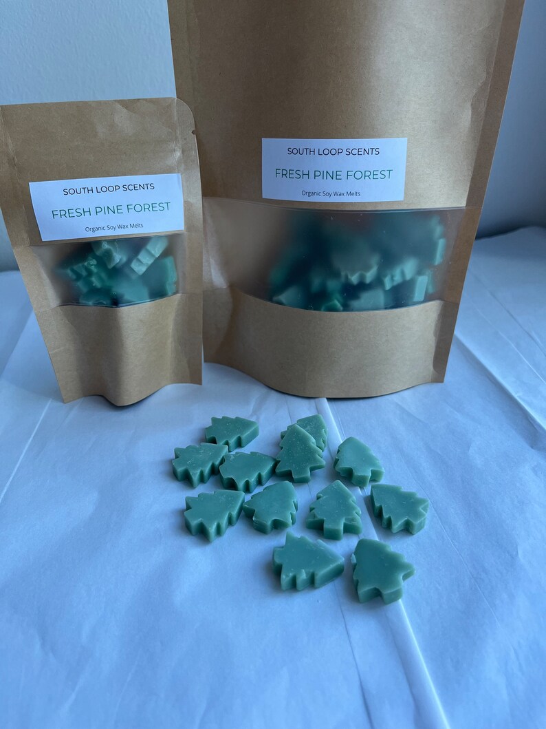 May include: Two brown paper bags with a clear window, each containing green wax melts shaped like pine trees. The bags have labels that read "South Loop Scents Fresh Pine Forest Organic Soy Wax Melts". A pile of green pine tree wax melts are scattered on a white surface.