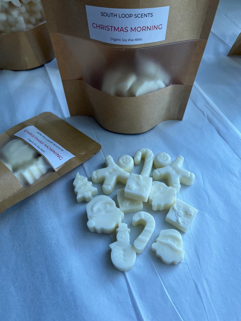 May include: A bag of white soy wax melts shaped like Christmas ornaments, including a snowman, a candy cane, a Christmas tree, and a Santa Claus. The bag is labelled "SOUTH LOOP SCENTS CHRISTMAS MORNING Organic Soy Wax Melts".