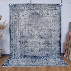 Puede incluir: Una alfombra rectangular grande con una apariencia vintage y desgastada. La alfombra presenta un diseño central en tonos grises y blancos, enmarcado por un borde azul oscuro. La alfombra se exhibe contra una pared gris claro.
