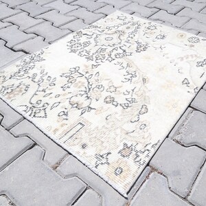 Puede incluir: Una alfombra blanca y beige con un diseño floral. La alfombra está colocada sobre una superficie de hormigón gris.