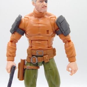 Op de afbeelding: Actiefiguur van een man met een ooglapje, gekleed in een oranje coltrui, groene broek en een bruine riem met holster. De figuur heeft zwarte schouderbeschermers en houdt een zwart pistool vast.