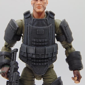 Op de afbeelding: Gedetailleerde actiefiguur van een soldaat, gekleed in een donkergrijs tactisch vest en bijpassende arm- en beenpantsering. De figuur houdt een zwart geweer vast. De soldaat heeft een geschilderd gezicht met camouflage-markeringen.