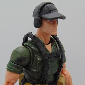 Może przedstawiać: Szczegółowa figurka żołnierza, nosząca ciemnoszarą czapkę z ochroną słuchu. Figurka ma zieloną koszulę w kamuflażu i czarną kamizelkę taktyczną. Odcień skóry figurki jest jasny, a tło jest gładkie.
