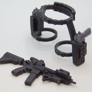 May include: Black toy rifle with scope and magazine, alongside a black tactical belt and holster set. The belt features two circular loops and a vertical support. The items are made of plastic.
