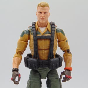 May include: Action figure of a male character with short blond hair, wearing a tan uniform with black tactical gear, green pants, and gloves. The figure has a serious expression and is posed against a plain background. The figure has a black belt and a black harness.