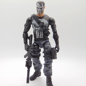 May include: A detailed action figure of a character in a tactical outfit, holding a rifle. The figure has a skull-like mask and is dressed in gray and black camouflage. The figure is standing on a white surface.