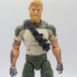 Op de afbeelding: Actiefiguur van een man met een blonde baard en haar, gekleed in een groen en wit shirt, een groene broek en een zwarte riem. De figuur heeft tatoeages op beide armen en grijze handschoenen.