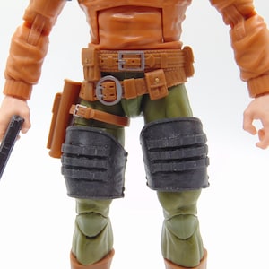 Op de afbeelding: Actiefiguur van een personage in een oranje jas, olijfgroene broek en bruine laarzen. De figuur heeft een bruine riem met zakjes, zwarte kniebeschermers en houdt een zwart pistool vast. De figuur is een verzamelobject.