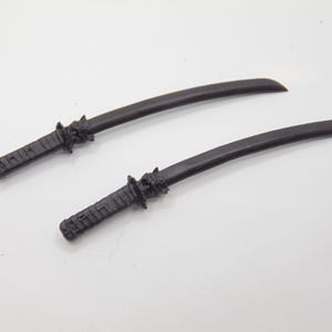 May include: Two black miniature katana swords. The blades are curved, and the handles are textured. The swords are lying on a white surface.