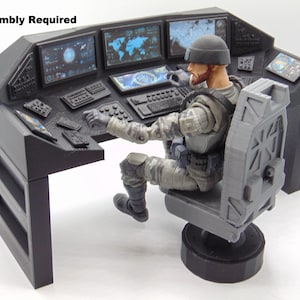 Könnte beinhalten: Ein grauer Kommandostand mit mehreren Bildschirmen, die Daten anzeigen, einer Tastatur und einer sitzenden Actionfigur in Militäruniform. Der Text "Assembly Required" ist sichtbar. Die Figur sitzt auf einem grauen Stuhl.