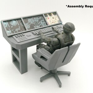 May include: A grey miniature command centre with a seated figure. The desk has three screens, a keyboard, and control panels. The figure is wearing a helmet and tactical gear. The text "Assembly Required" is visible.
